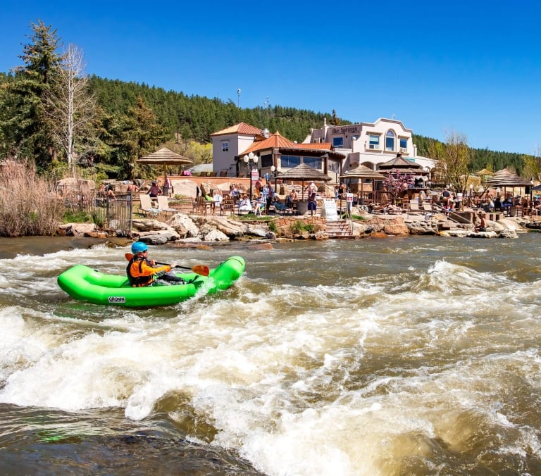 A person in a green CRONIN inflatable kayak paddles through rapids. 'The Springs' resort with people and umbrellas lines the bank, backed by a forested hill.