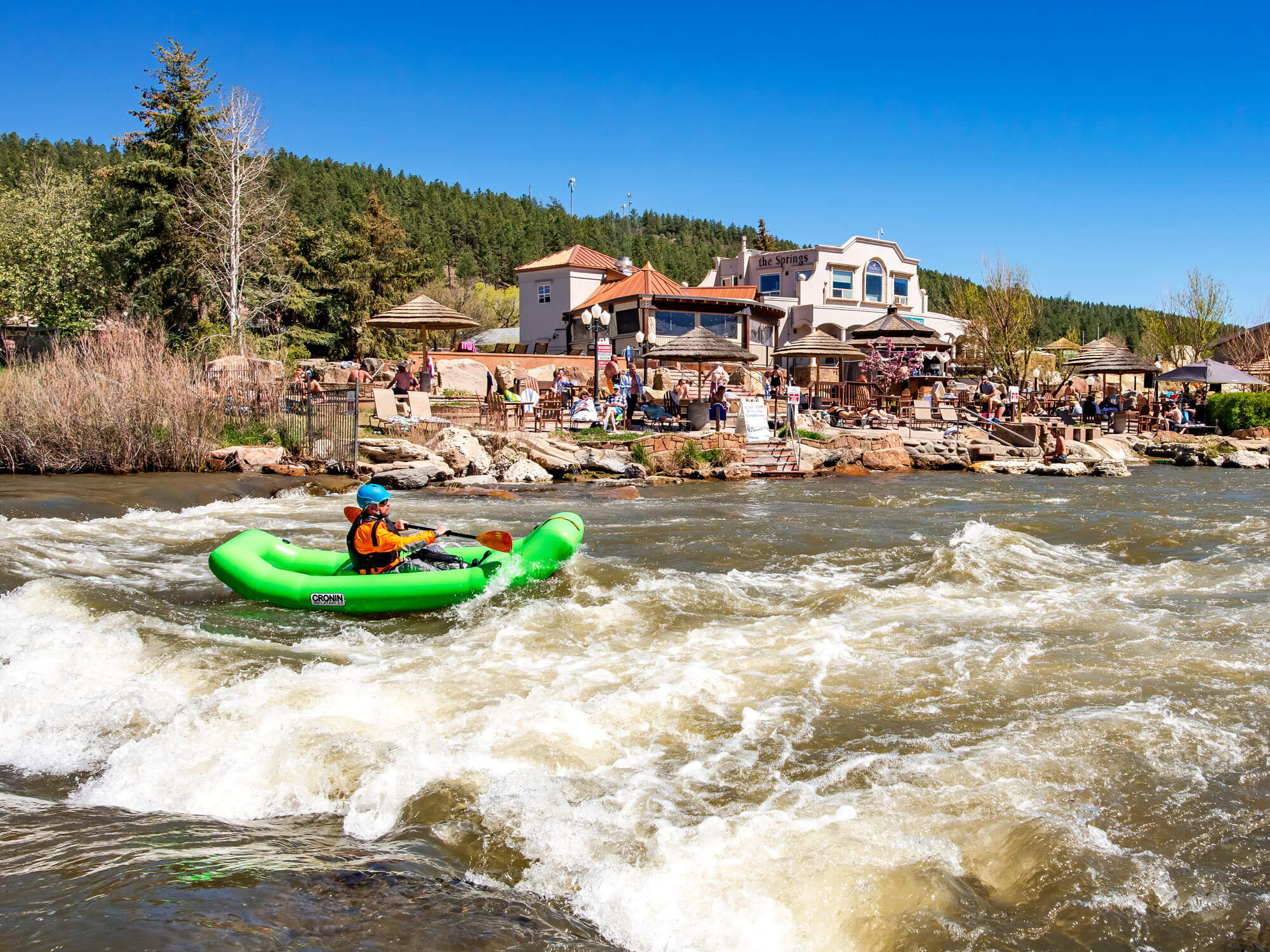 A person in a green CRONIN inflatable kayak paddles through rapids. 'The Springs' resort with people and umbrellas lines the bank, backed by a forested hill.