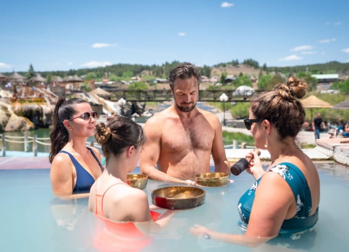 Four people are immersed in a light blue outdoor pool, arranging three golden singing bowls. One woman holds a mallet. This takes place at a resort under a bright blue sky.
