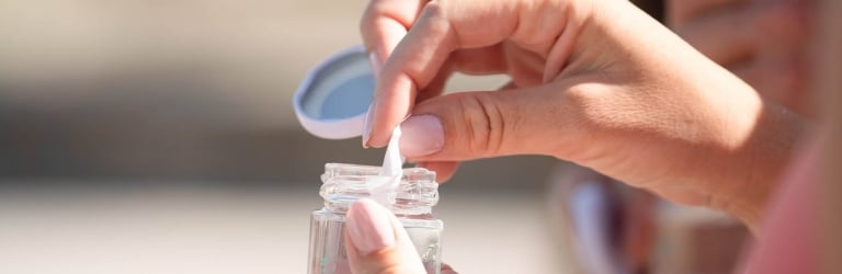 A person's hands open a small clear glass jar, pulling a white applicator from the liquid within, set against a blurred outdoor background.
