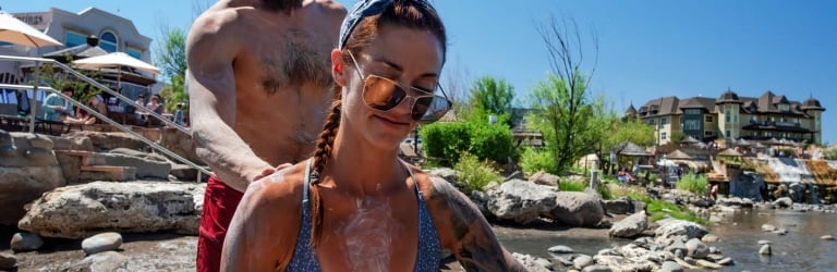 Woman and man, covered in white mud, apply it to each other by a muddy riverbank. 