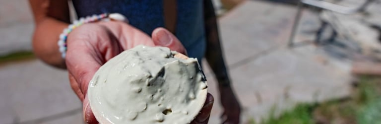 A hand holds an open clam shell filled with light grey mud. A person in a blue swimsuit stands by water, rocks, and grass.
