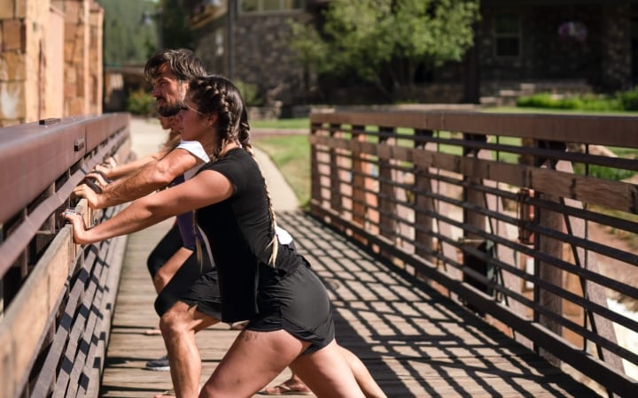 A woman with braided hair and a man are stretching, leaning forward against a wooden railing. They are on a sunny wooden bridge with buildings and greenery in the background.
