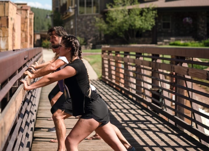 A woman with braided hair and a man are stretching, leaning forward against a wooden railing. They are on a sunny wooden bridge with buildings and greenery in the background.