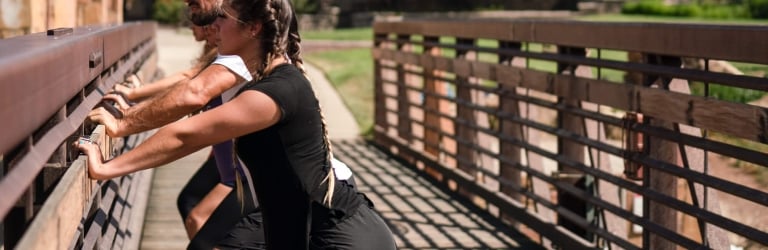 A woman with braided hair and a man are stretching, leaning forward against a wooden railing. They are on a sunny wooden bridge with buildings and greenery in the background.