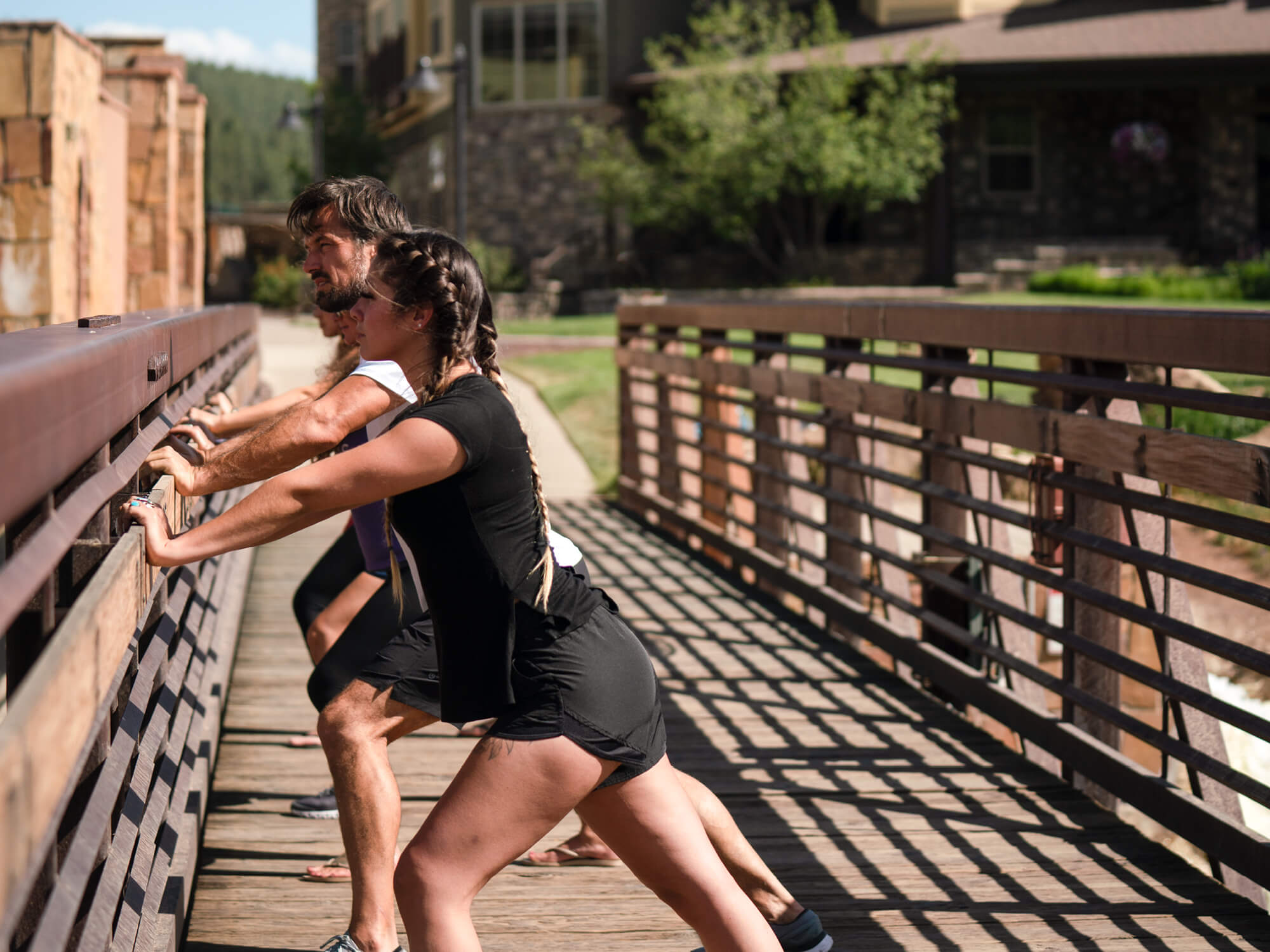 A woman with braided hair and a man are stretching, leaning forward against a wooden railing. They are on a sunny wooden bridge with buildings and greenery in the background.