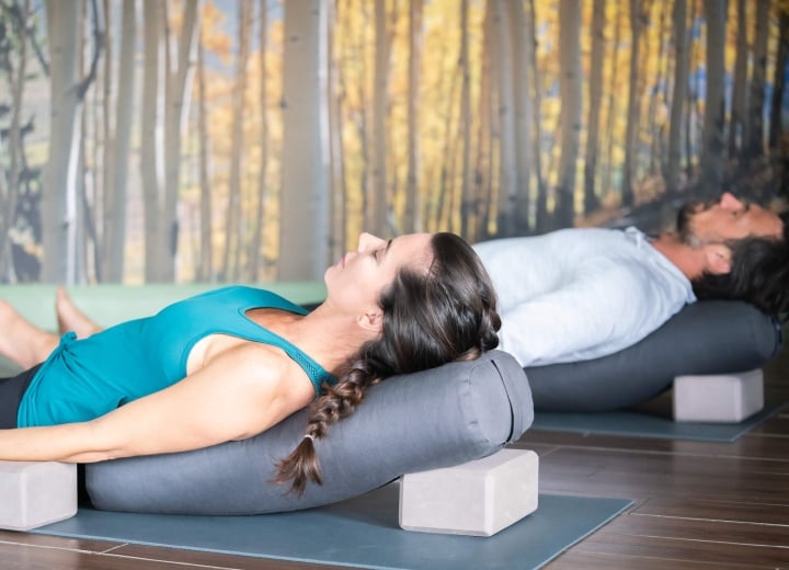A woman and man lie relaxed on their backs with eyes closed, supported by grey bolsters and blocks on yoga mats, in a room with a birch forest mural.
