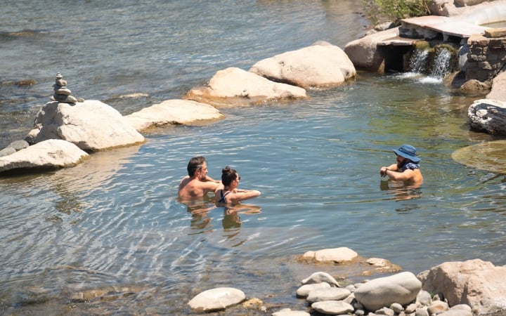 Three people relax chest-deep in a natural river hot spring, surrounded by rocks and a small man-made waterfall, under bright daylight.