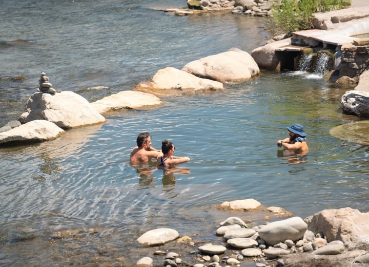 Three people relax chest-deep in a natural river hot spring, surrounded by rocks and a small man-made waterfall, under bright daylight.