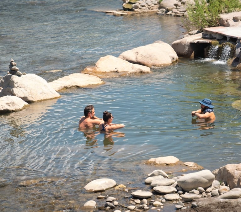 Three people relax chest-deep in a natural river hot spring, surrounded by rocks and a small man-made waterfall, under bright daylight.