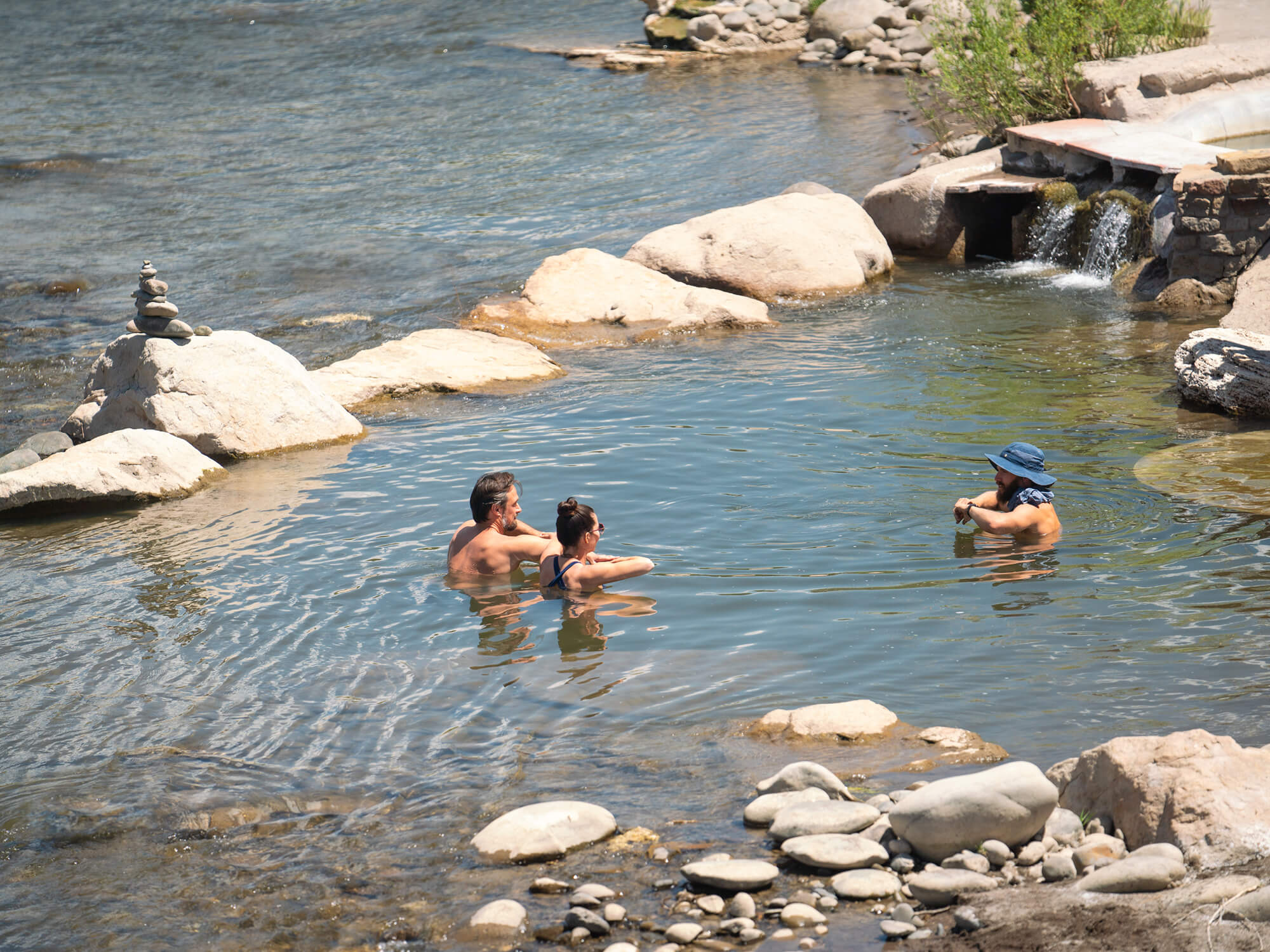 Three people relax chest-deep in a natural river hot spring, surrounded by rocks and a small man-made waterfall, under bright daylight.