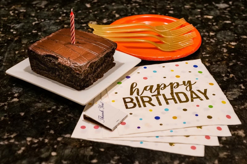 A chocolate cake slice with a red-striped candle is displayed on a white plate. Nearby are napkins featuring 