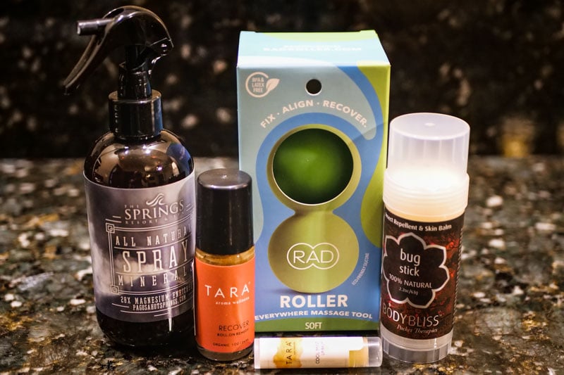 Various wellness and personal care products are displayed on a dark, speckled countertop. Text: THE SPRINGS RESORT ALL NATURAL SPRAY MINERAL 20X MAGNESIUM ENRICHED PAGOSAHOTSPRINGS.COM; TARA aroma wellness™ RECOVER ROLL ON REMEDY ORGANIC .33 OZ / 10 ML; BPA & LATEX FREE FIX · ALIGN · RECOVER. RAD ROLLER EVERYWHERE MASSAGE TOOL SOFT ECO FRIENDLY SILICONE; TARA COOL REFRESH LIP BALM; Insect Repellent & Skin Balm bug stick 100% NATURAL 2.2oz / 63g BODY BLISS Pocket Therapies™.