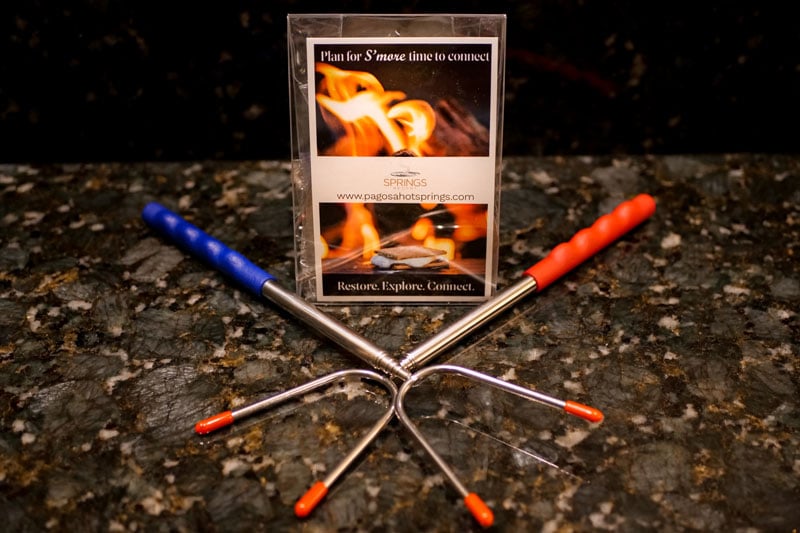 Two metal telescoping s'mores roasting sticks, with blue and red handles, are crossed on a dark granite counter. A promotional card stands behind them, displaying: 