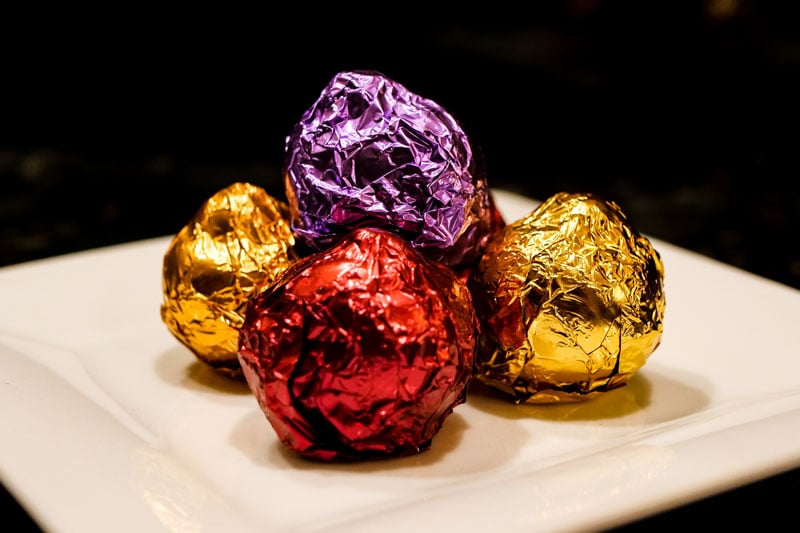 Four chocolates wrapped in crumpled red, purple, and gold foil are piled on a white rectangular plate against a dark background.