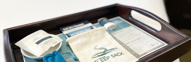 A dark wooden tray presents hotel amenity items: a white sachet, a blue pouch, and a 