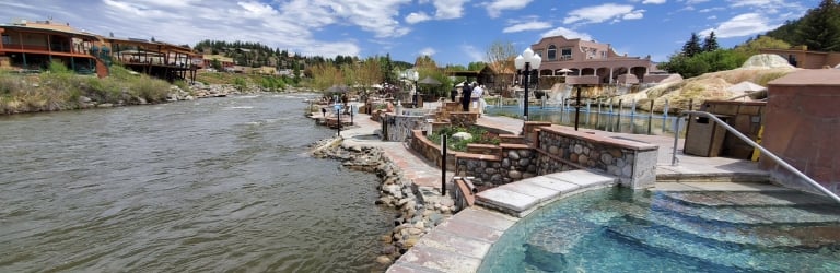 A large outdoor hot springs complex with several turquoise pools and people relaxing, set along a wide, swift river. Buildings and hills line the far bank under a partly cloudy sky.
