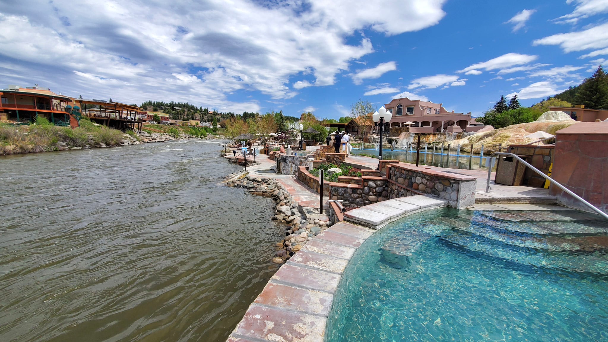 A large outdoor hot springs complex with several turquoise pools and people relaxing, set along a wide, swift river. Buildings and hills line the far bank under a partly cloudy sky.
