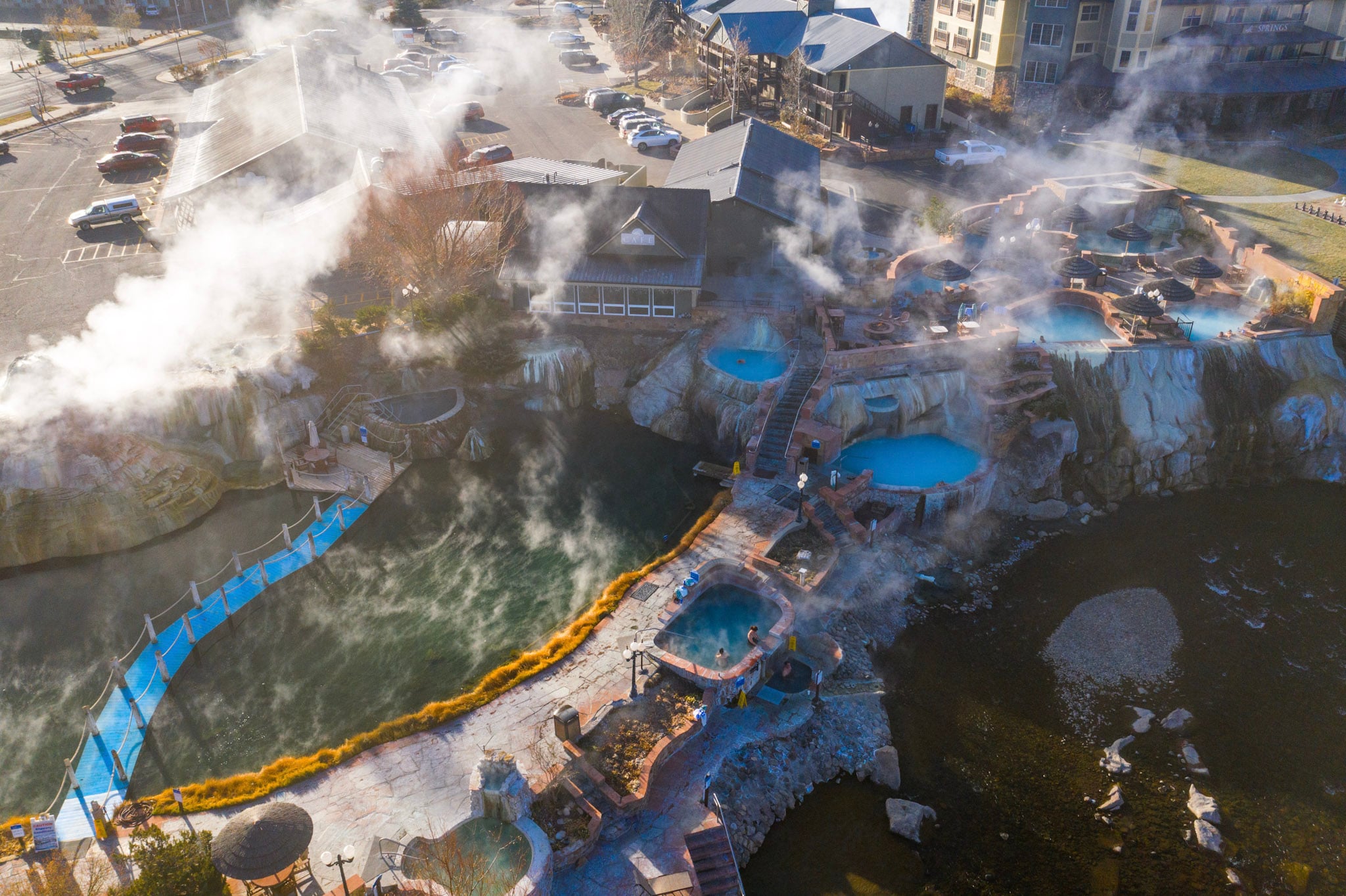 Multiple steaming hot spring pools are tiered along a river, connected by pathways and a blue bridge with people soaking in the water. Nearby are urban buildings, including a 