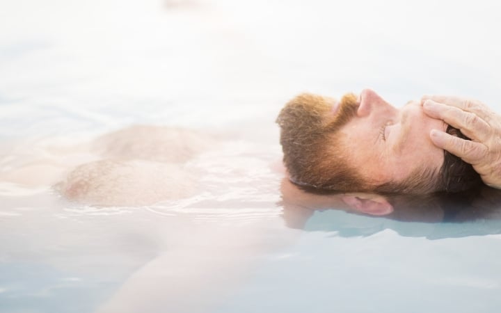 A red-bearded man floats on his back, eyes closed, with his right hand resting on his forehead, partially submerged in light blue, cloudy water.