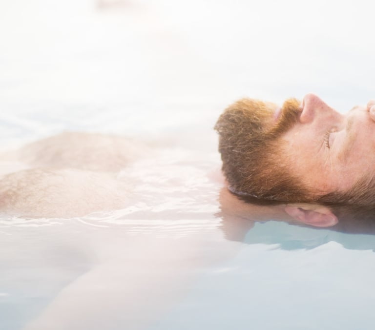 A red-bearded man floats on his back, eyes closed, with his right hand resting on his forehead, partially submerged in light blue, cloudy water.