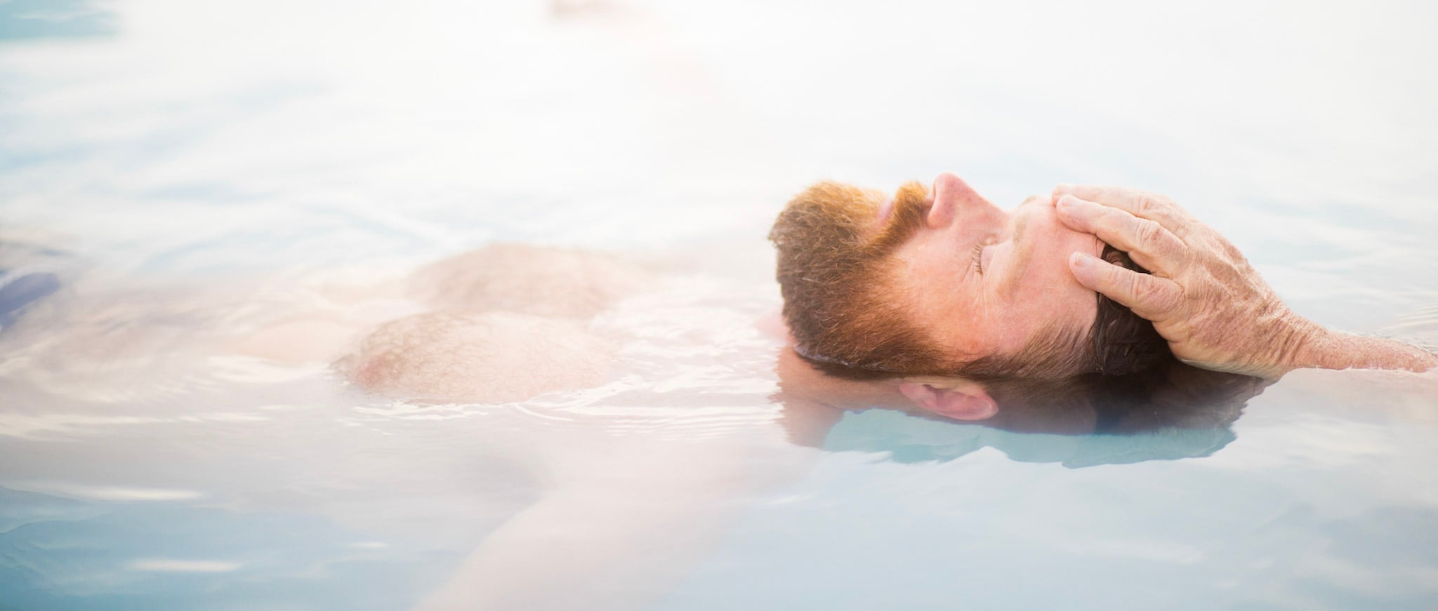 A red-bearded man floats on his back, eyes closed, with his right hand resting on his forehead, partially submerged in light blue, cloudy water.