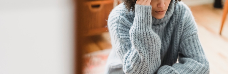 A woman with curly dark hair sits on the floor, pressing her hand to her forehead, looking pensive. She wears a blue knit sweater and jeans in a room with blurred wooden furniture.