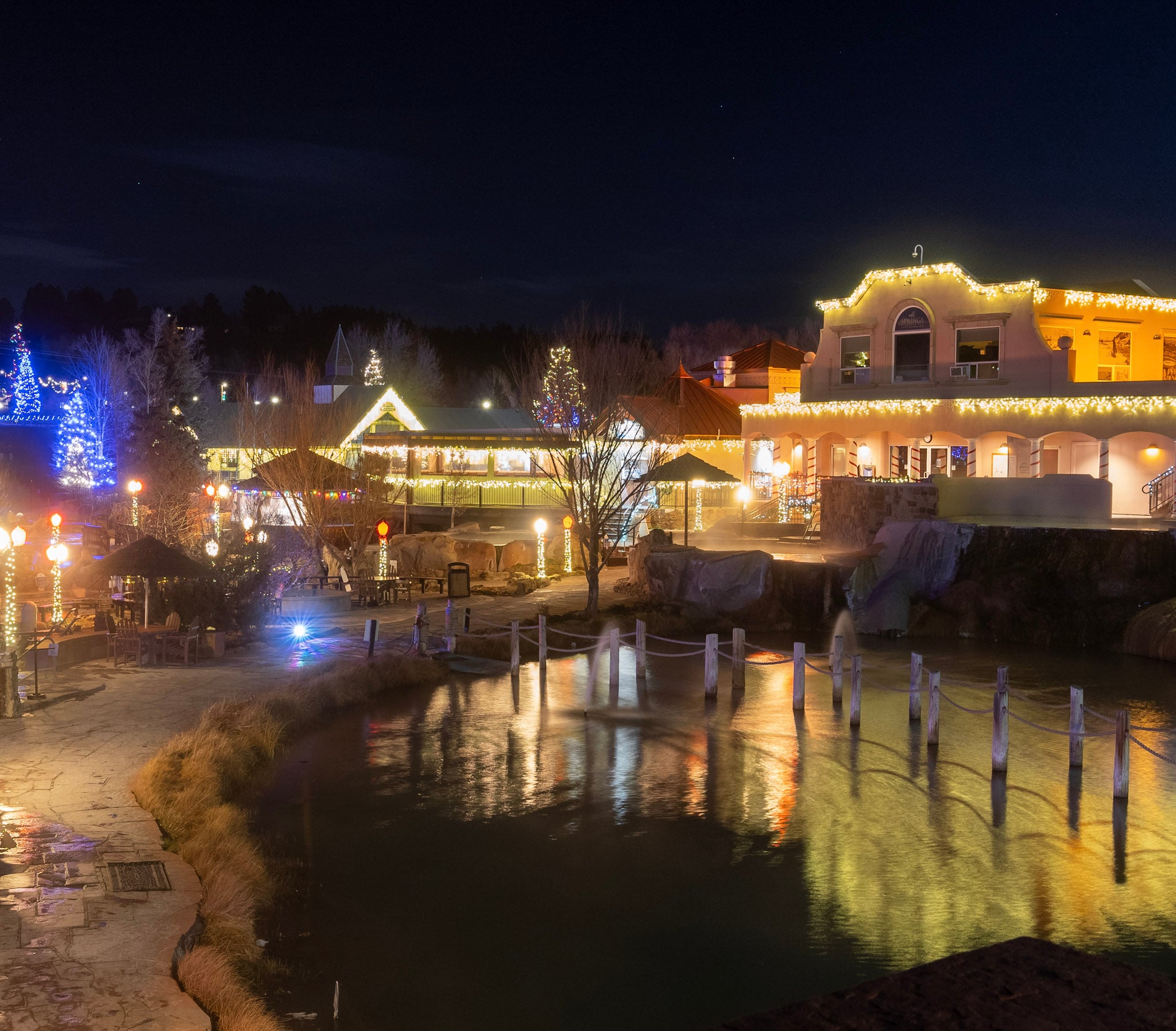 A resort complex, illuminated by festive yellow and blue lights, reflects in a wide pond. 'THE SPRINGS' is on the main building, all under a dark night sky.