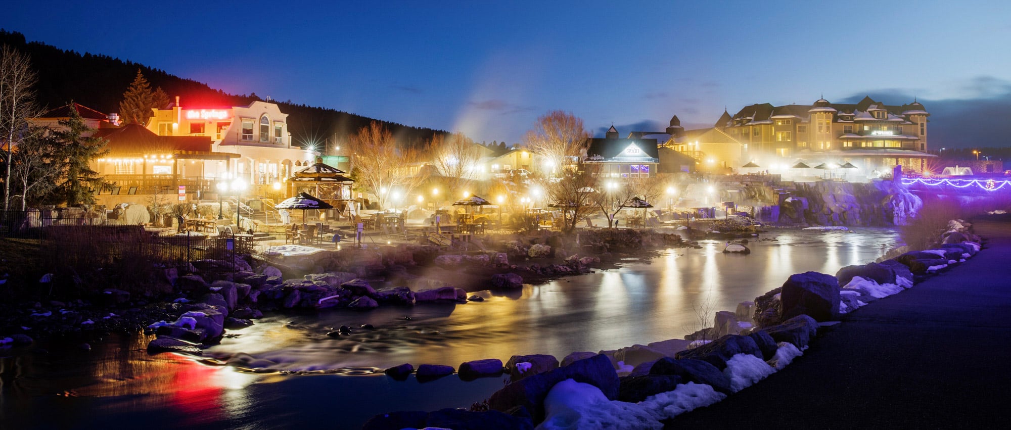 A winter hot springs resort glows brightly with steaming pools and illuminated buildings lining a river, against a dark forested hillside. Text: Hot Springs