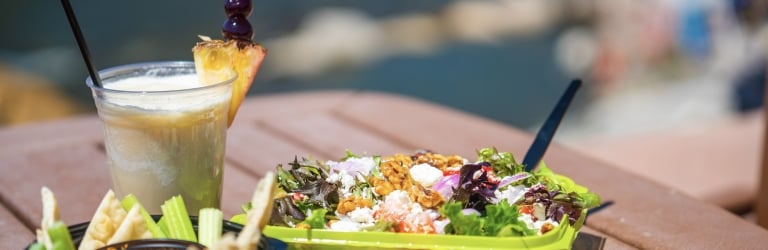 A tropical drink with fruit garnish, a vibrant salad, and a dip with pita and celery are displayed on a brown table near blurred water.