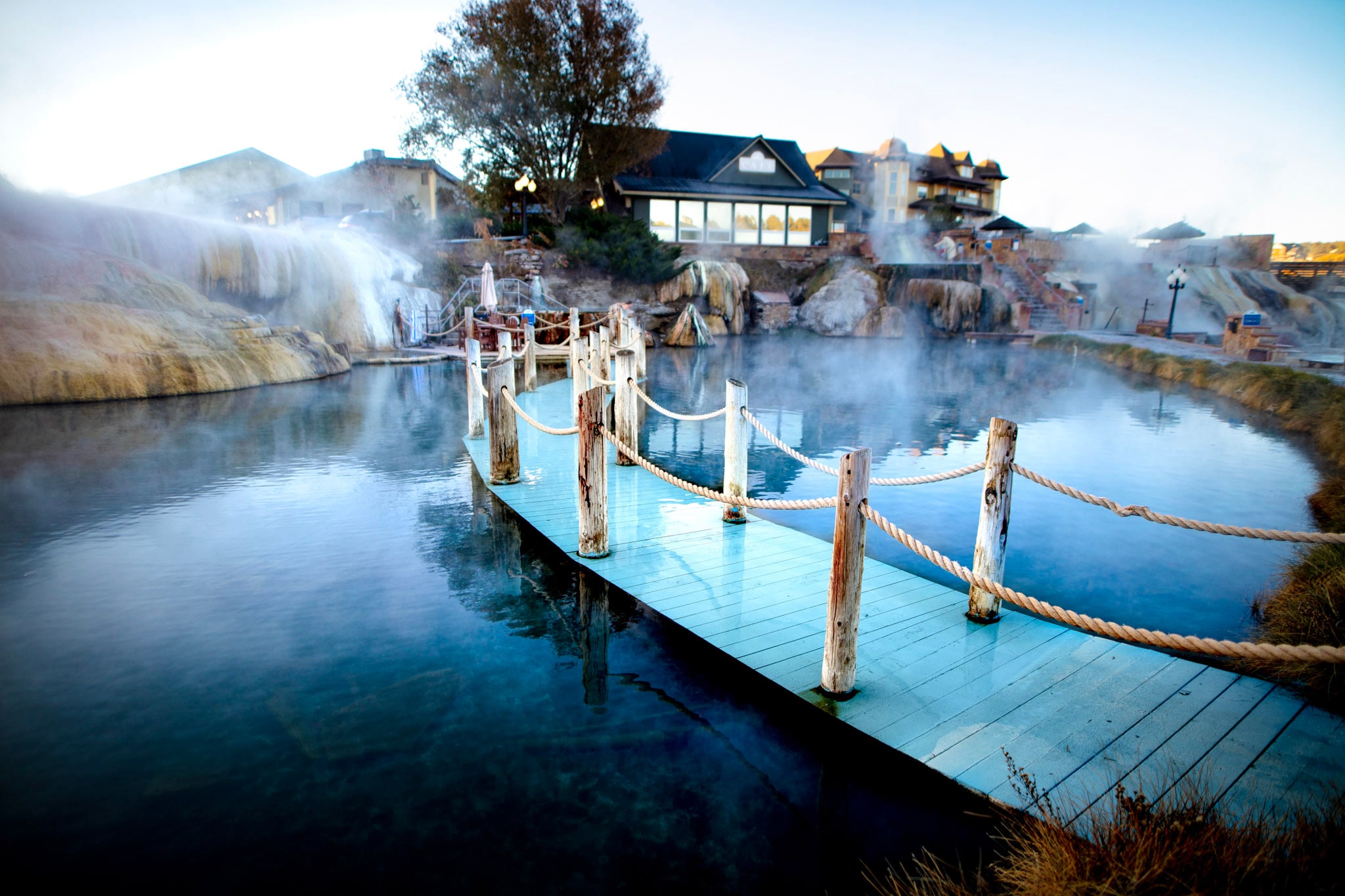 A light blue wooden pier stretches over steamy, blue-green water. In the background, resort buildings and steam rise from multi-tiered, mineral-stained hot spring formations.