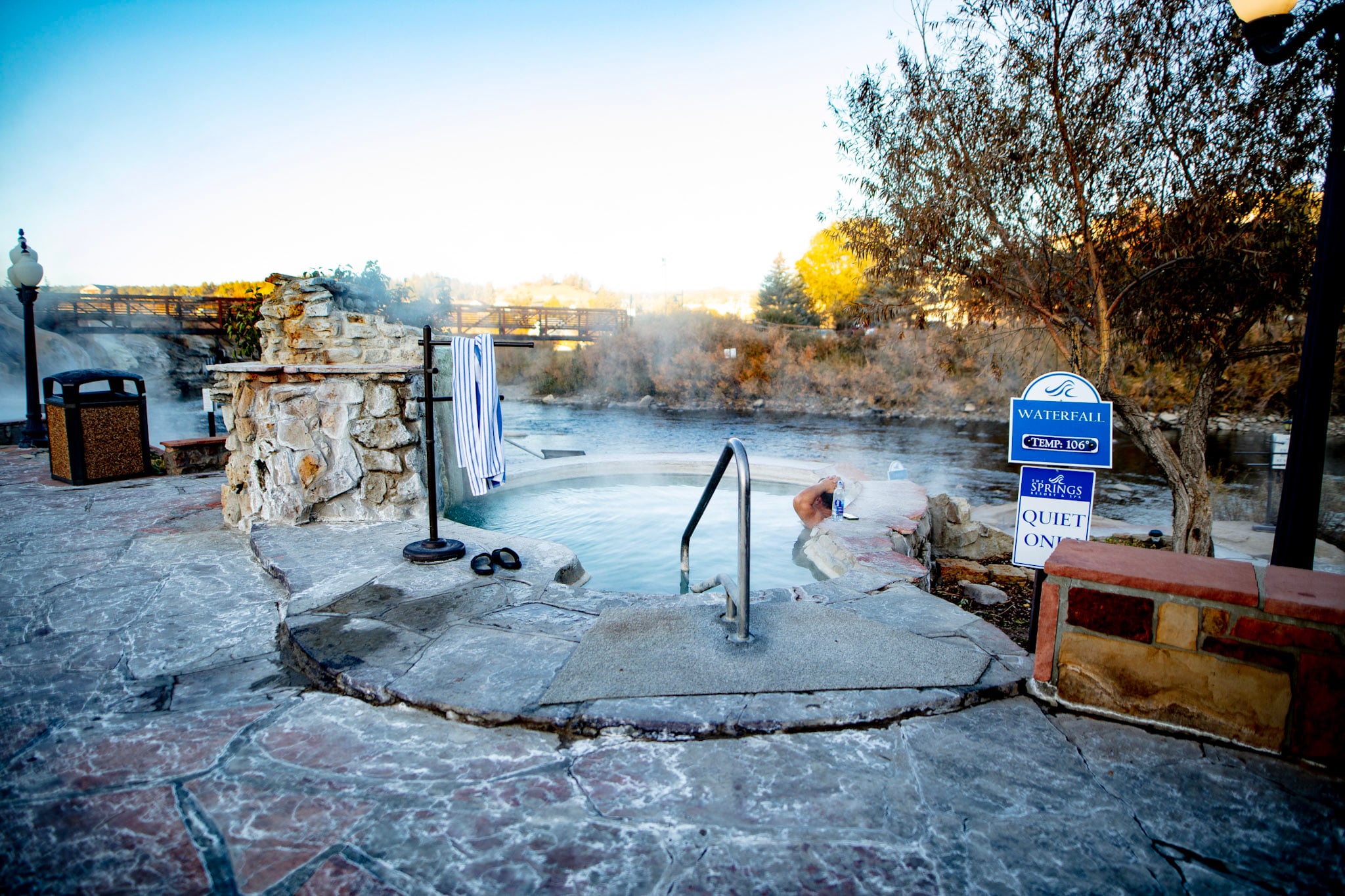 A steaming outdoor hot tub with a person relaxing, flanked by stone walls and a river, with a distant bridge under a clear sky. Signs read: WATERFALL TEMP: 106° THE SPRINGS RESORT & SPA QUIET ON!