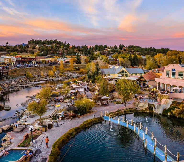 Outdoor hot spring pools, busy with people, line a river. A town (featuring 
