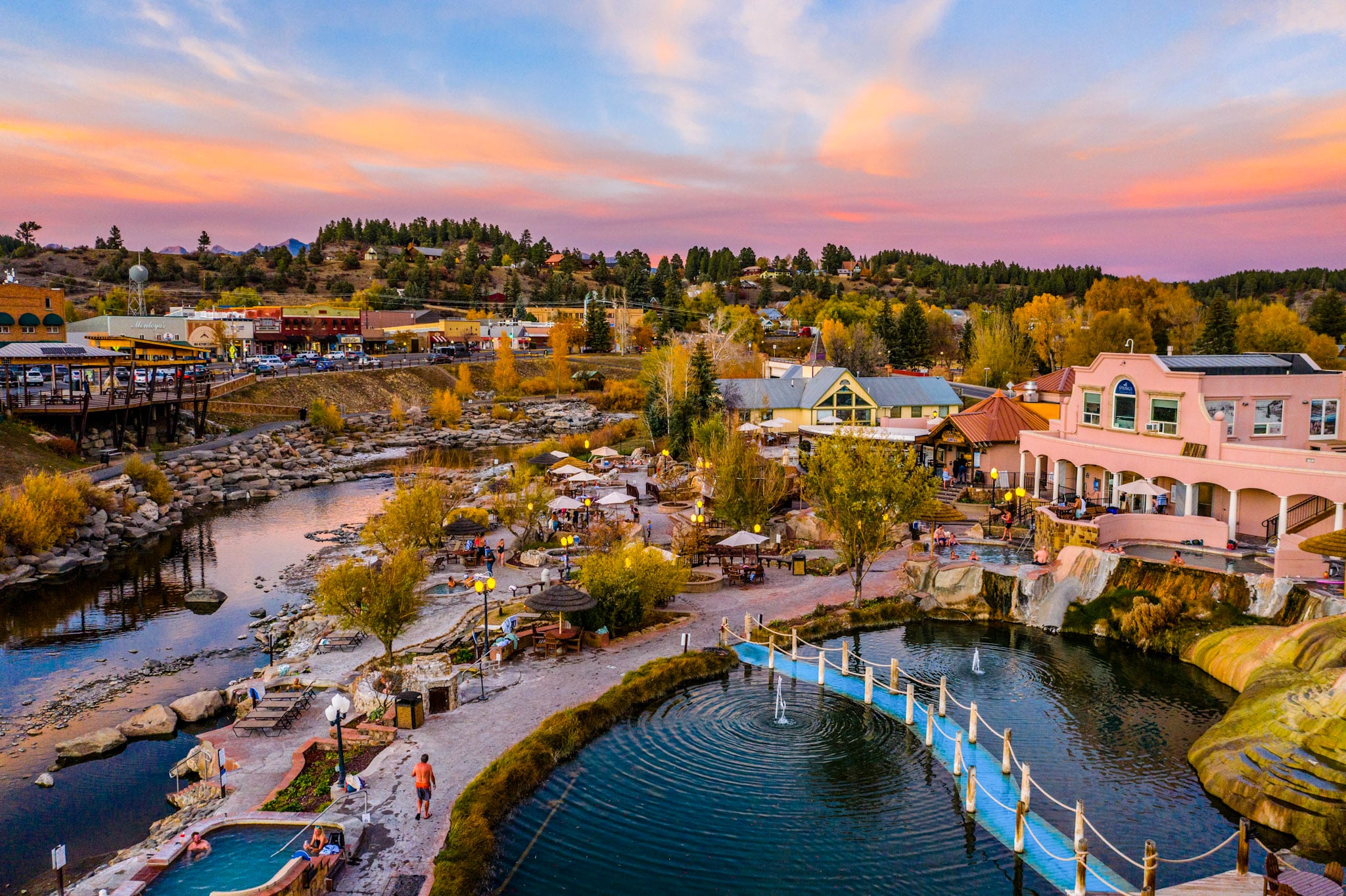 Outdoor hot spring pools, busy with people, line a river. A town (featuring 