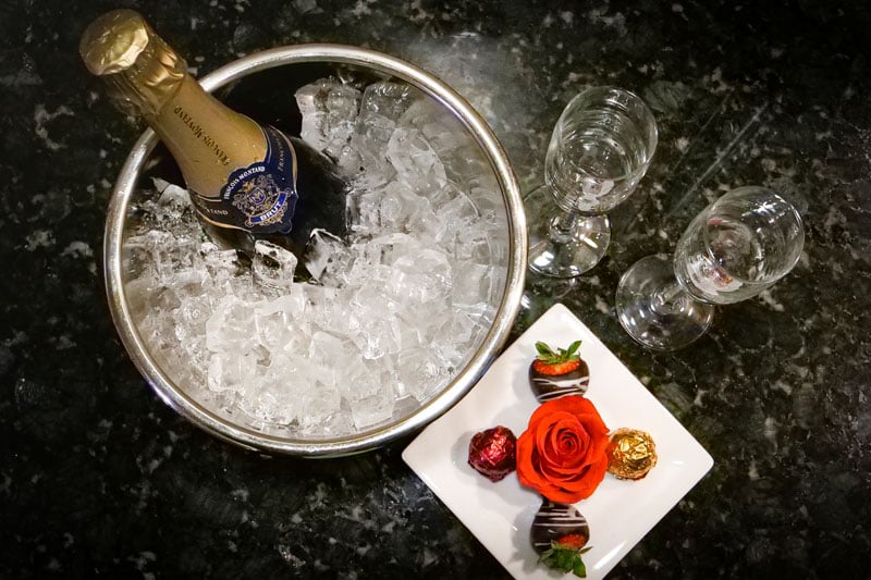 A champagne bottle chilling in an ice bucket. Two empty glasses and a white plate with a red rose, chocolate-dipped strawberries, and wrapped chocolates rest on a dark granite counter. The champagne bottle label reads: FRANCOIS MONTAND, BLANC DE BLANCS, BRUT, METHODE TRADITIONNELLE, PRODUCT OF FRANCE.
