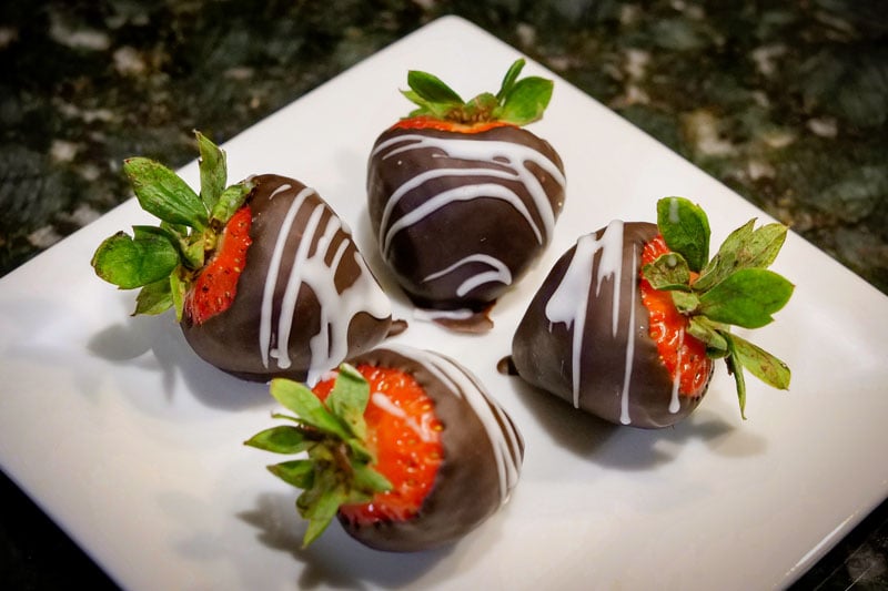 Four chocolate-covered strawberries, featuring dark chocolate with white drizzles, are arranged on a white square plate against a dark, mottled background.