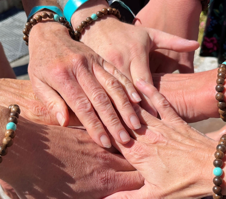 Several hands, adorned with brown beaded bracelets featuring turquoise beads and turquoise wristbands, are stacked together in a huddle, bathed in bright sunlight.