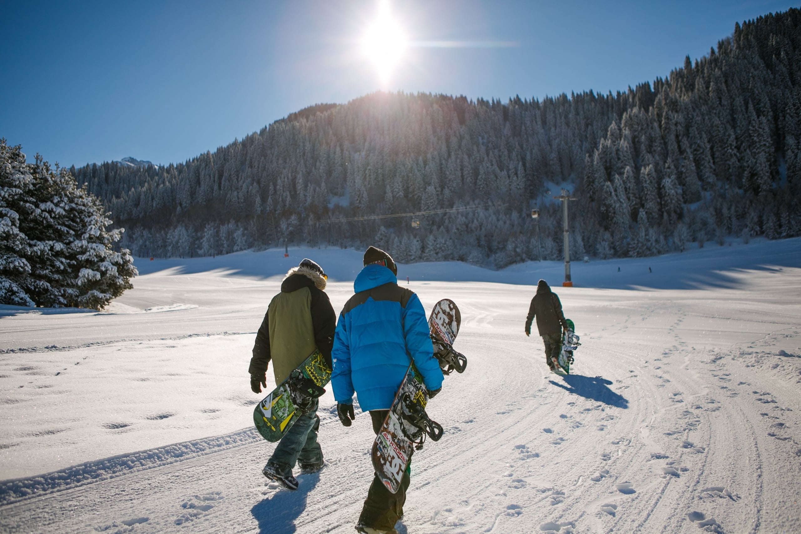 Three snowboarders walk away on a groomed snowy slope towards sunlit evergreen mountains with ski lifts. Snowboards show 