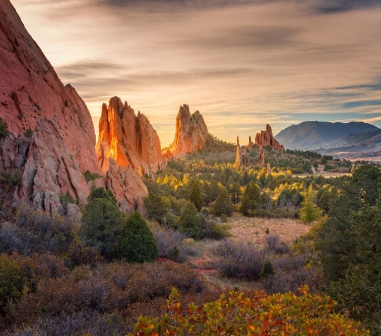 Towering red rock formations stand amidst vibrant autumn foliage in a valley, illuminated by warm sunrise light under a cloudy sky with distant mountains.
