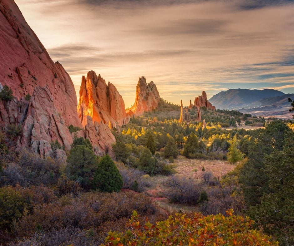 Towering red rock formations stand amidst vibrant autumn foliage in a valley, illuminated by warm sunrise light under a cloudy sky with distant mountains.
