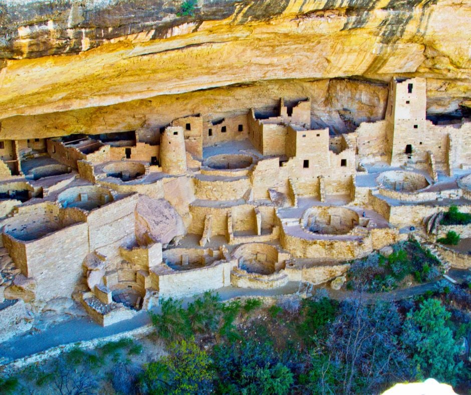 Ancient pueblo cliff dwellings, with stone rooms and circular kivas, sit preserved under a vast golden rock overhang, above a verdant valley.