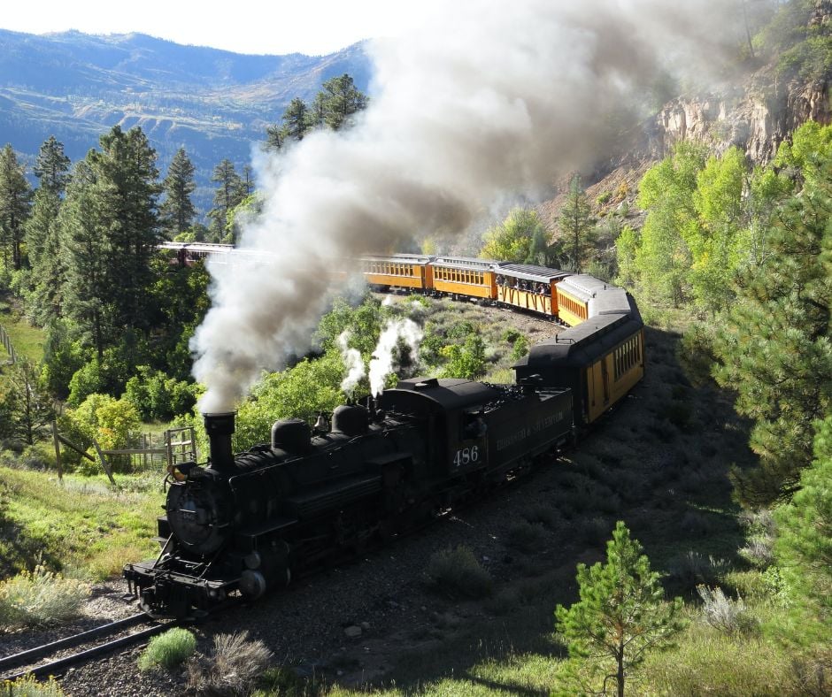 A black steam locomotive 486, DURANGO & SILVERTON, emits thick smoke while pulling yellow passenger cars around a curve through a lush, green mountainous landscape with pine trees and cliffs.