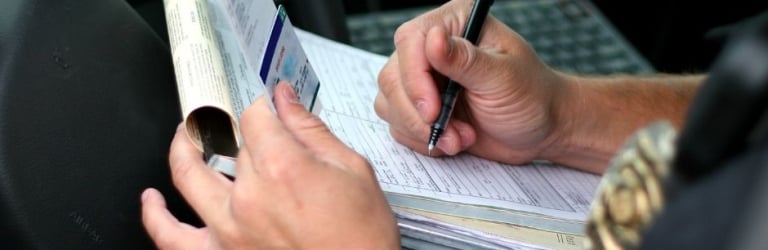 An officer's hands write on a form with a pen, holding a rolled document and card, inside a vehicle with an 