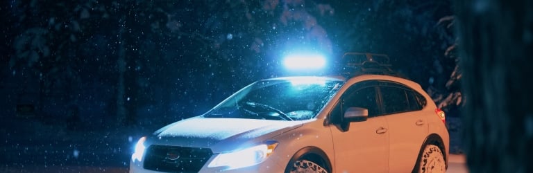 A white Subaru crossover SUV with a roof light bar and headlights on is parked, covered in fresh snow, during a dark, heavy snowfall in a forest.