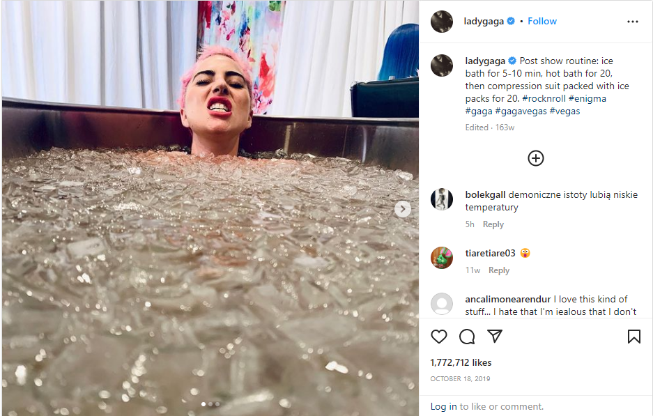 Lady Gaga, with pink hair and a grimacing expression, is submerged in a brown tub filled with ice and water, against a white curtain and colorful artwork.