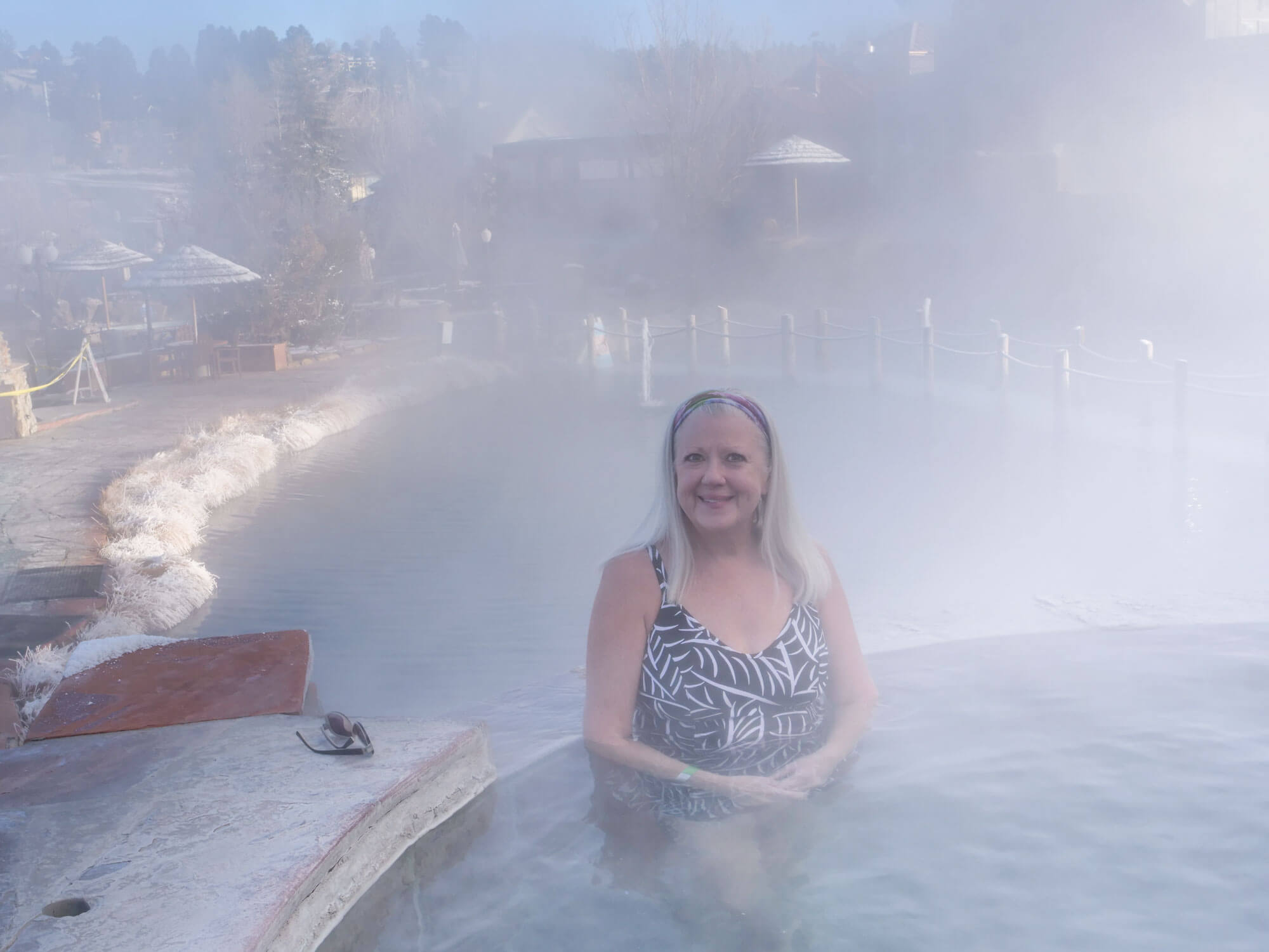 A smiling woman with white hair and a patterned swimsuit is partially submerged in a very steamy outdoor hot spring, with snow-covered banks and bare trees visible through the mist.