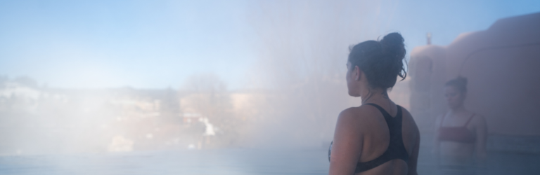 A woman in a black swimsuit stands in a steamy outdoor pool, looking left. A second woman, distant landscape, and reddish building are visible under a blue sky.