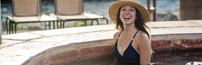 A woman in a straw hat and swimsuit smiles while soaking in an outdoor hot tub, with lounge chairs and water visible in the background.