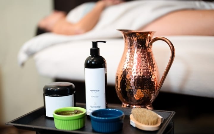 A black tray holds a pump bottle, jar, two small bowls, a brush, and a hammered copper pitcher, set before a person on a massage bed.
Text: HERBAL BODY OIL Rejuvenating Action For all skin types With Almond & Apricot Kernel Oil for all skin types FOR PROFESSIONAL USE