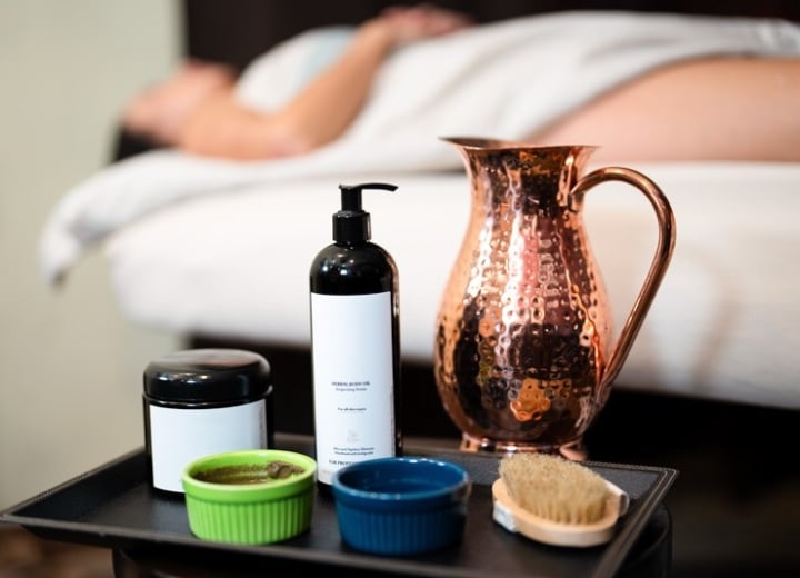 A black tray holds a pump bottle, jar, two small bowls, a brush, and a hammered copper pitcher, set before a person on a massage bed.
Text: HERBAL BODY OIL Rejuvenating Action For all skin types With Almond & Apricot Kernel Oil for all skin types FOR PROFESSIONAL USE